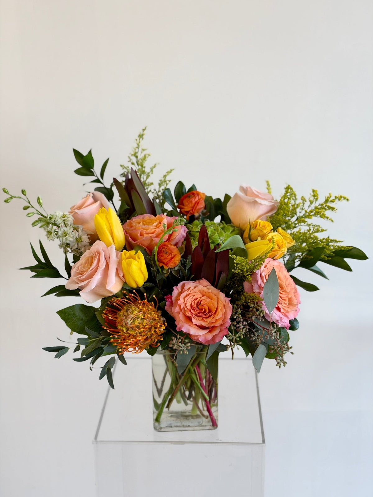 Sunset arrangement of peach roses, yellow tulips and an orange pincushion protea in a glass vase