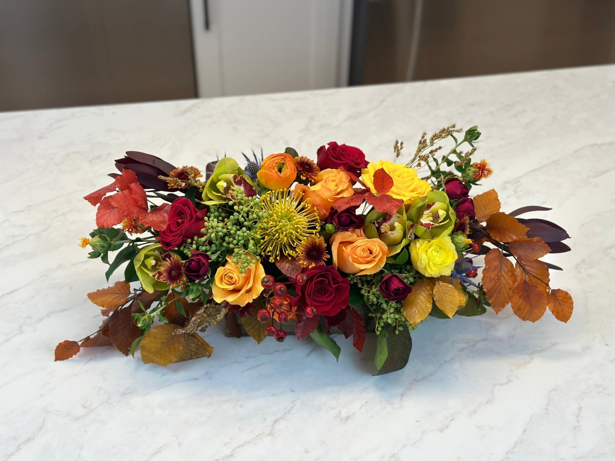 Long low fall centerpiece of red, orange and yellow roses with autumn leaves on a marble table
