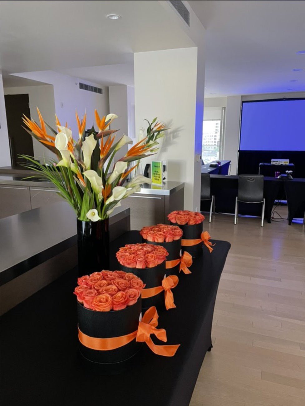 Orange roses in black hat boxes tied with ribbon beside a bird of paradise and calla arrangement, prepared as client gifts