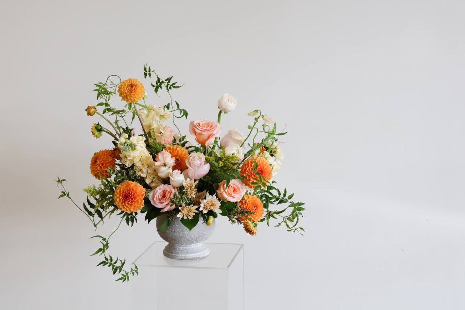 Compote arrangement of orange dahlias, peach garden roses and white ranunculus with trailing greenery on a white pedestal, designed by Novapetal in New York