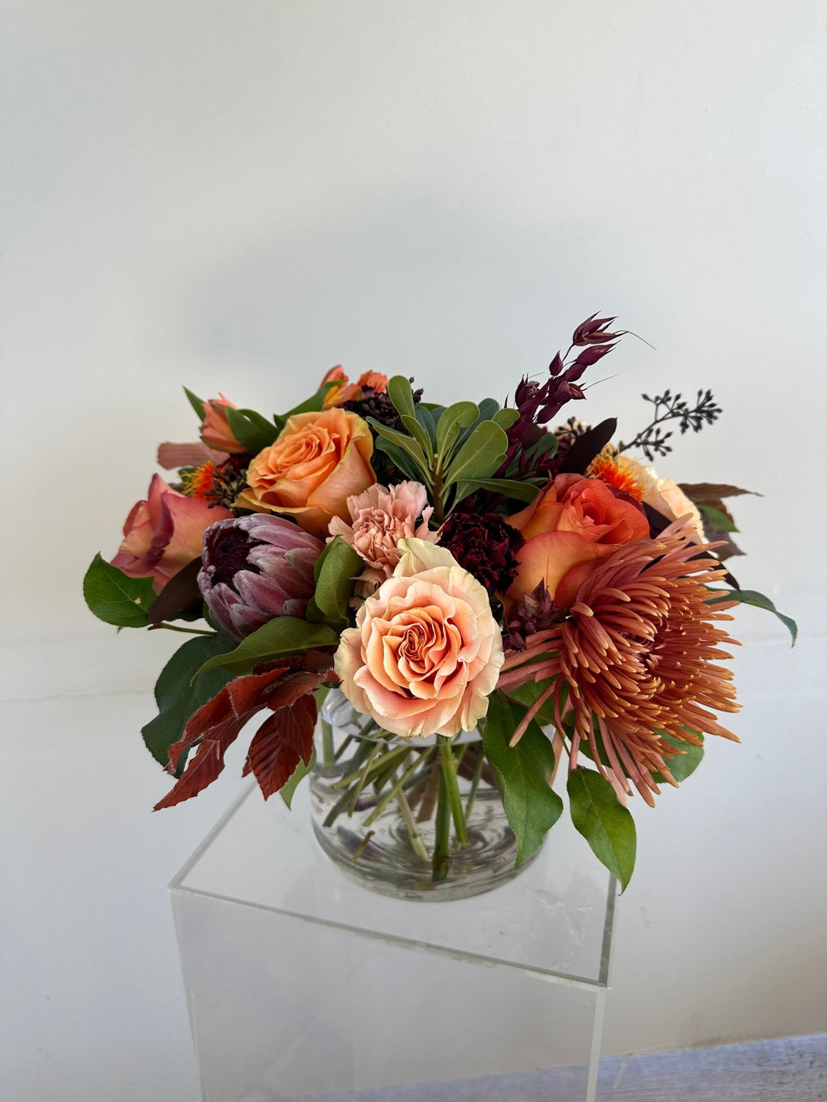 Autumn arrangement of peach and orange roses, a protea and a rust spider chrysanthemum in a round glass vase