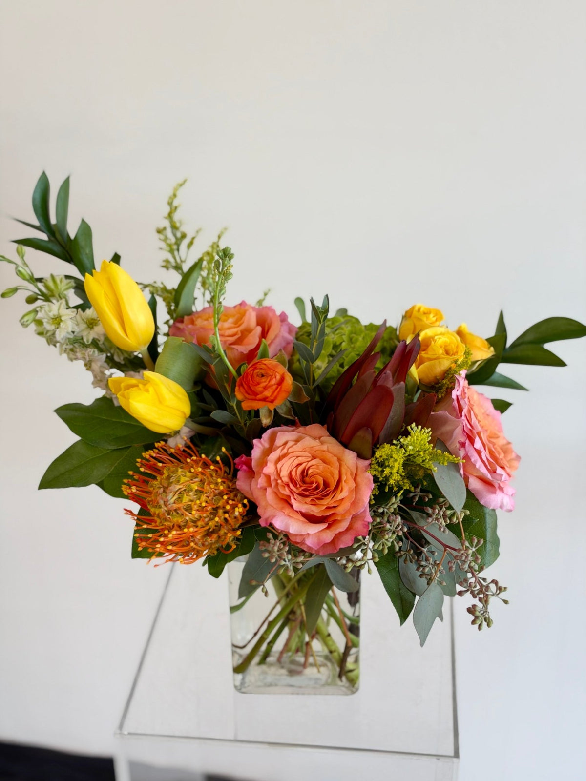Sunset arrangement of coral roses, yellow tulips and an orange pincushion protea in a rectangular glass vase