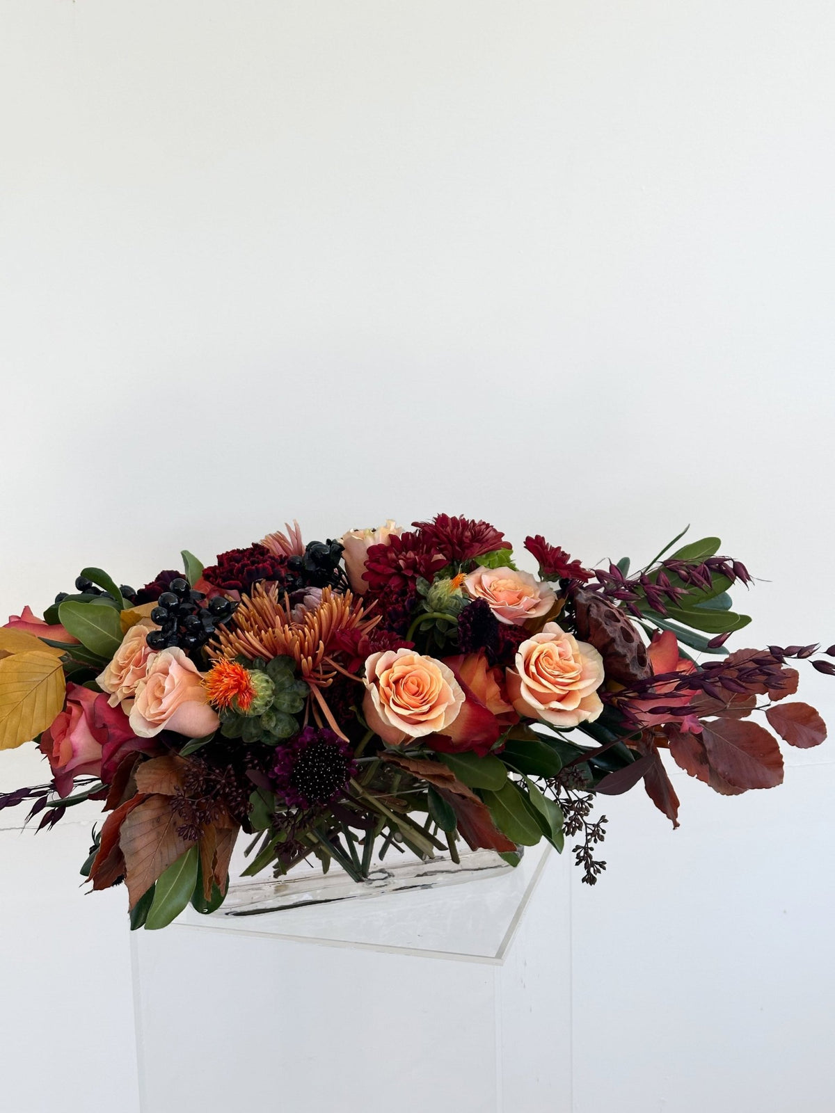 Long low autumn centerpiece of peach roses, burgundy and rust chrysanthemums and dark berries on a clear acrylic base