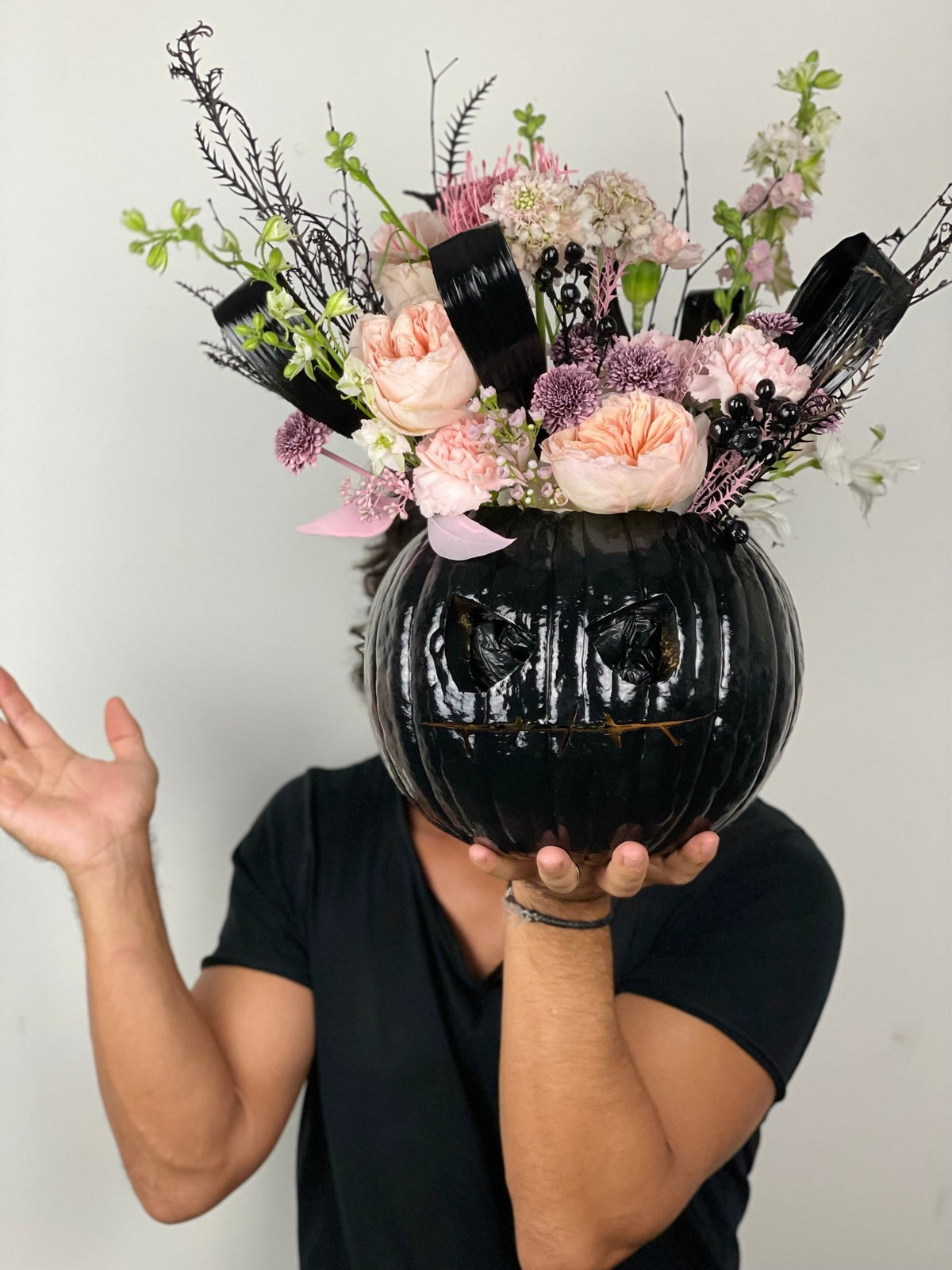 Person holding the Jack-o'-Lantern, a carved black pumpkin with peach garden roses, pink carnations and dark branches