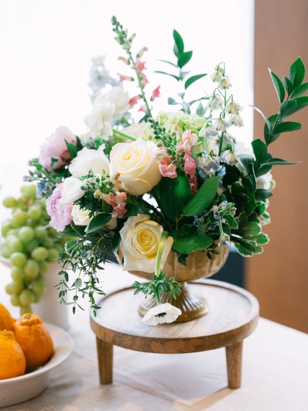 Pastel compote arrangement of garden roses, peonies, ranunculus and snapdragon on a wooden riser beside grapes and oranges