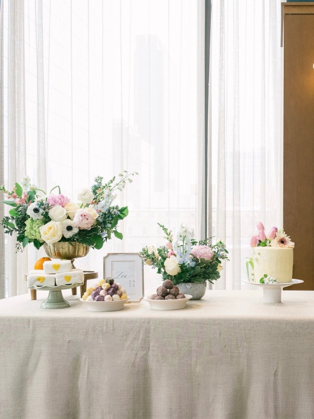 Baby shower table against a bright New York window with two pastel arrangements, a floral cake and pastel desserts
