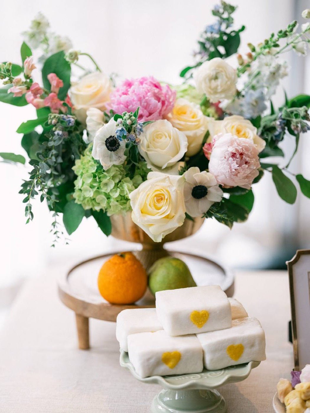 Pastel compote arrangement of peonies, garden roses, anemones and green hydrangea above a stand of heart-marked desserts