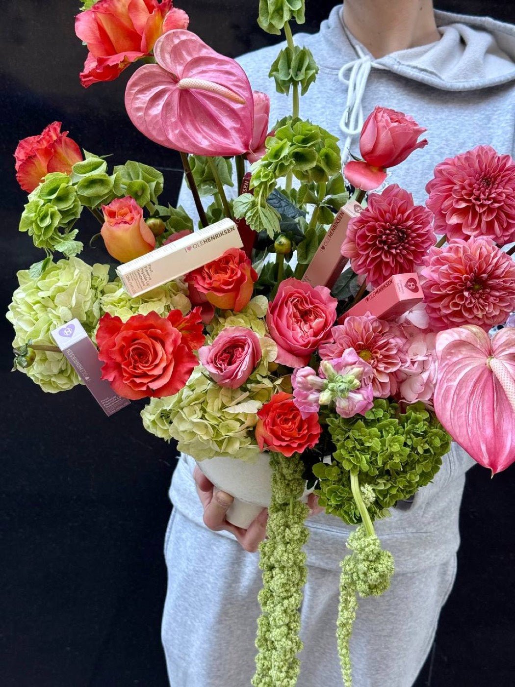 Gifting arrangement of coral roses, pink dahlias, anthurium and green hydrangea with a beauty brand's products set among the stems