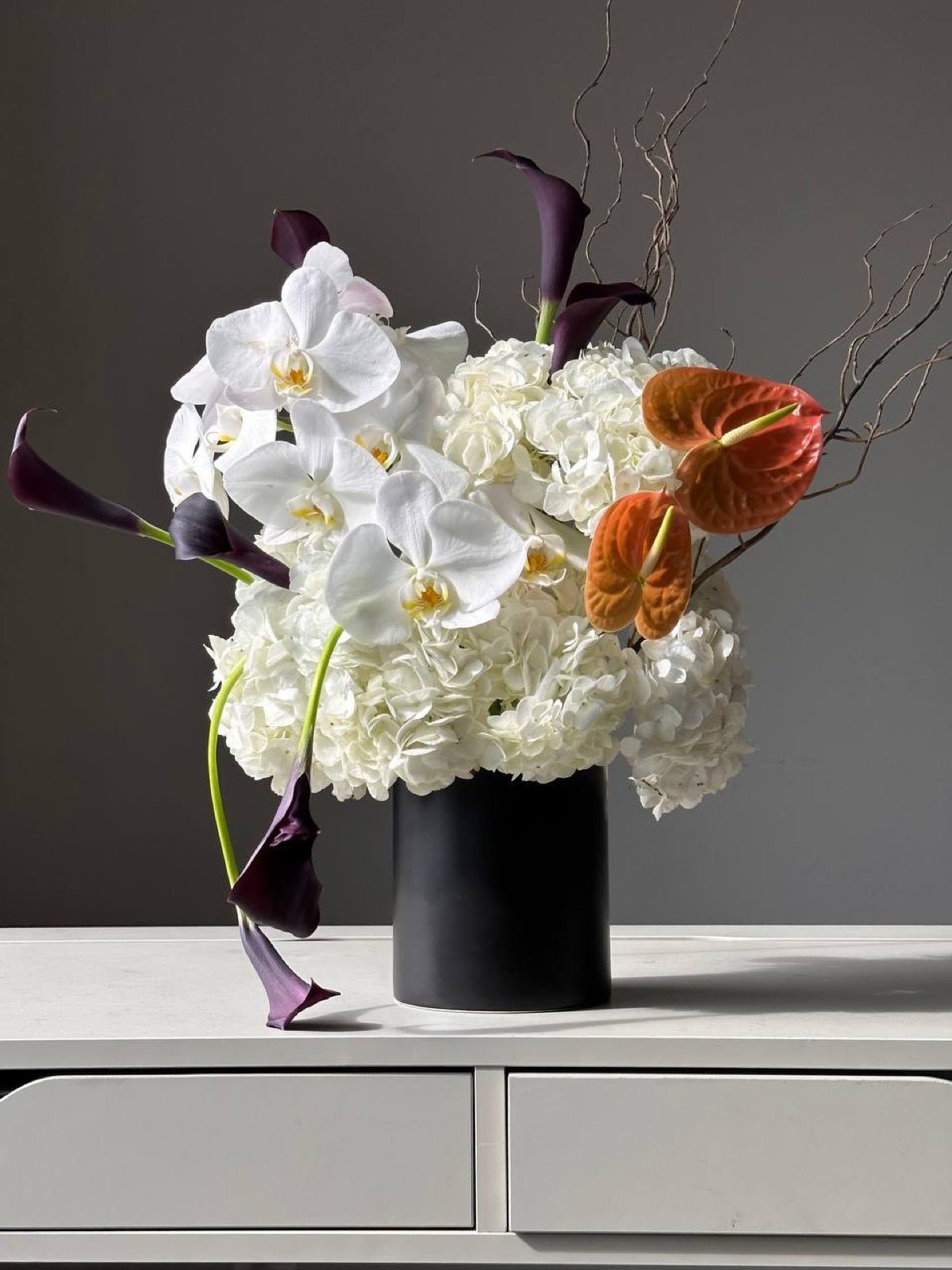 White hydrangea and phalaenopsis orchid arrangement with plum calla lilies and orange anthurium in a matte black vase, designed by Novapetal in New York