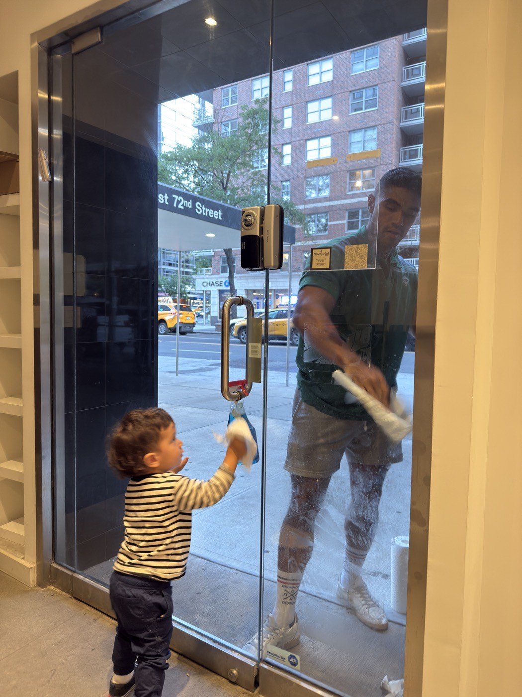 A small child helping wipe the glass door of the Novapetal studio on East 72nd Street while Anthony Sheppard cleans the glass from the street side