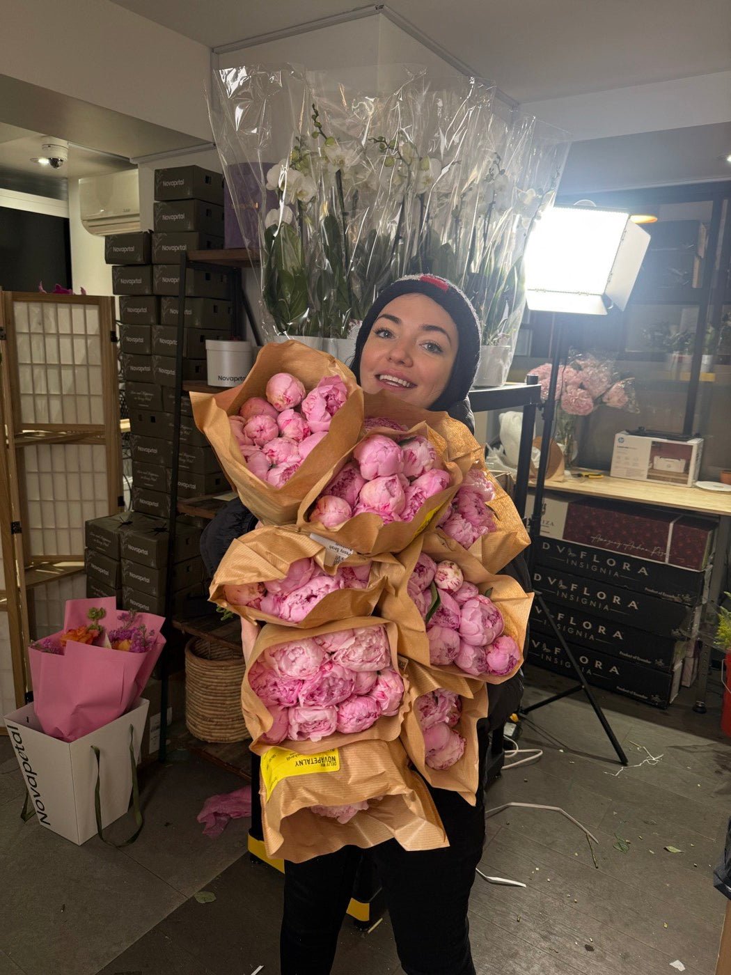 Olga Sheppard holding an armful of pink peonies in kraft wrap in the Novapetal studio, beside stacked Novapetal boxes and grower crates