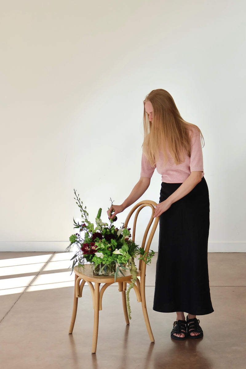 Novapetal designer finishing a burgundy and green arrangement on a bentwood chair