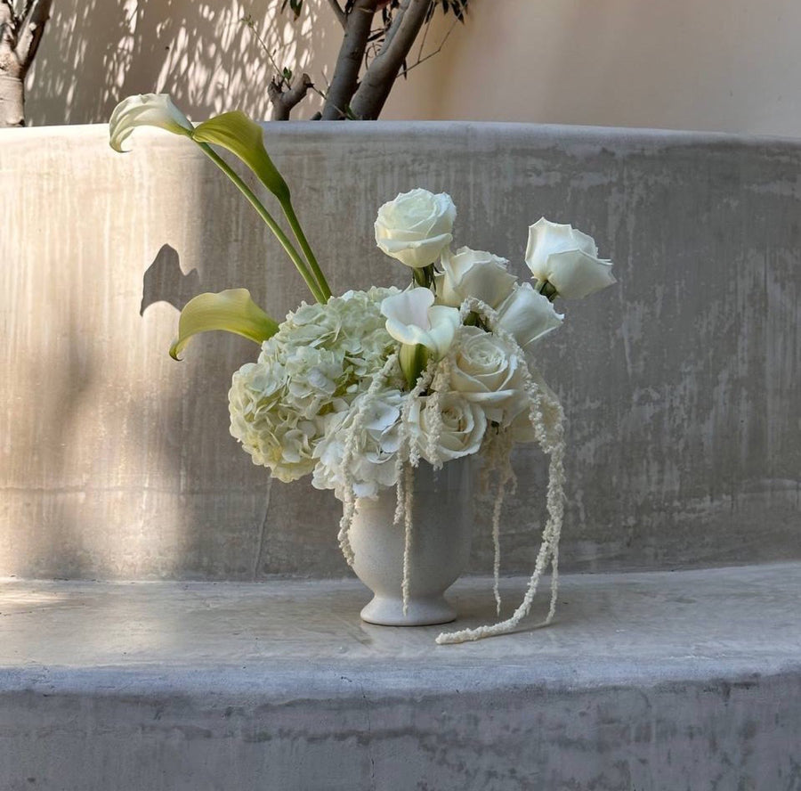 White calla lilies roses and hydrangea in a stone footed vase against a plaster wall