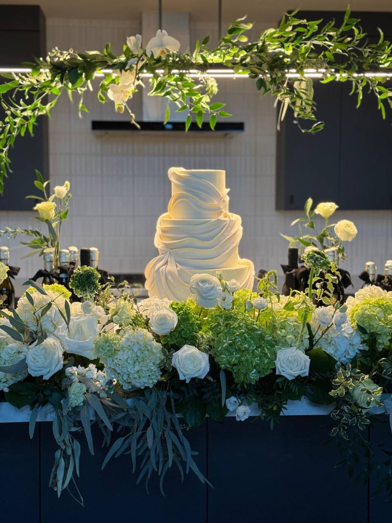 Wedding cake framed by a hanging greenery installation and a white flower bar